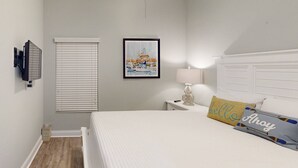 A bedroom with a bed, a bedside table with a lamp, a framed picture on the wall, and a window with blinds.