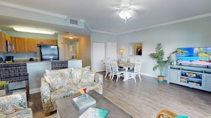 A living room with a sofa, coffee table, and a dining area with chairs. There is a kitchen in the background with wooden cabinets and a refrigerator.