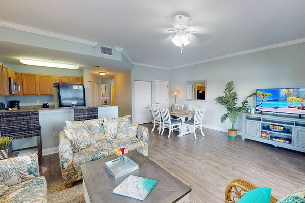 A living room with a sofa, coffee table, and a dining area with chairs. There is a kitchen in the background with wooden cabinets and a refrigerator.
