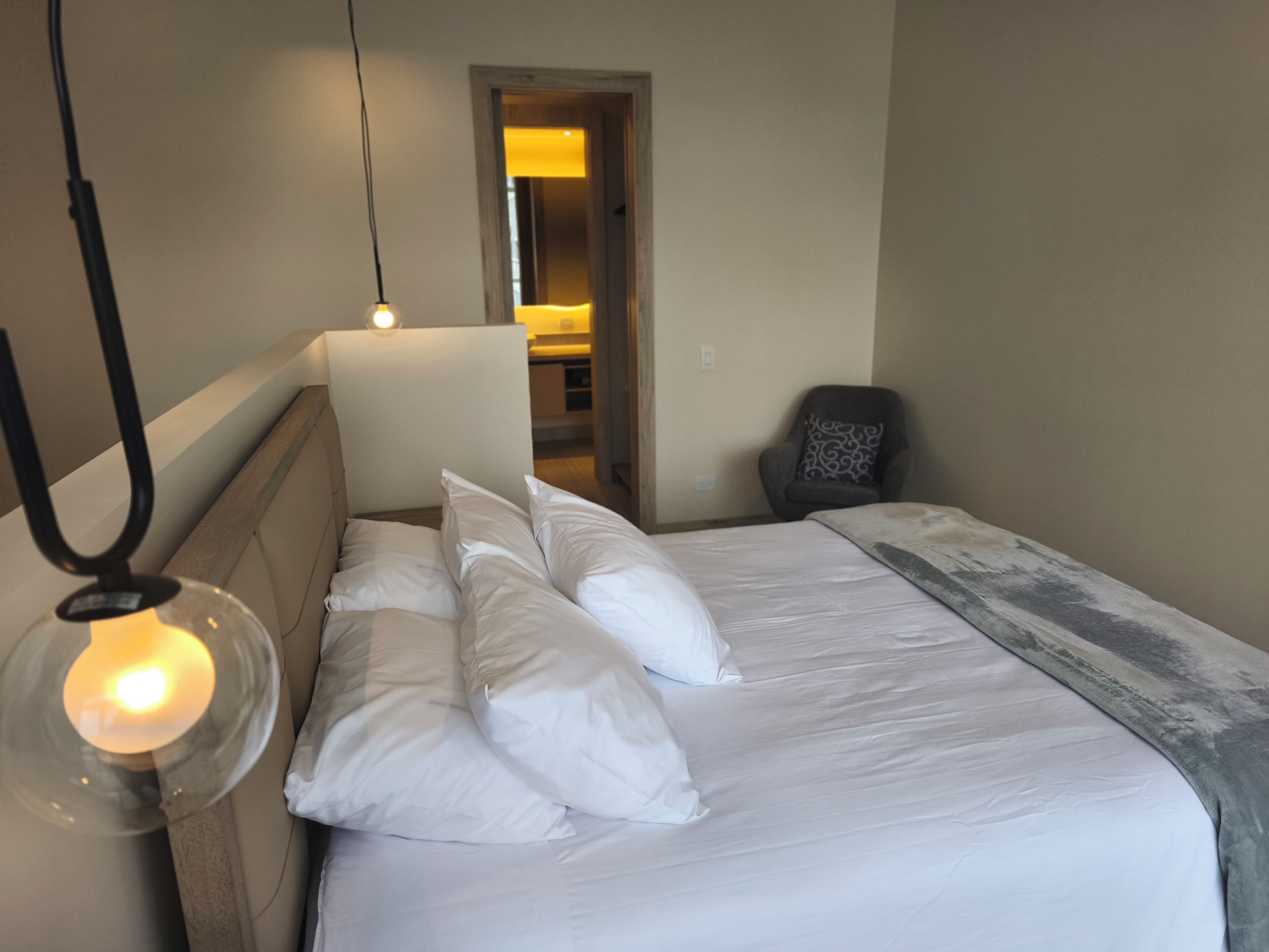 A neatly made bed with white linens and pillows, a bedside lamp, a chair with a patterned cushion, and a doorway leading to a bathroom.