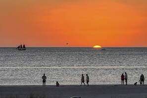 A sunset over a beach with people walking and a sailboat in the distance.