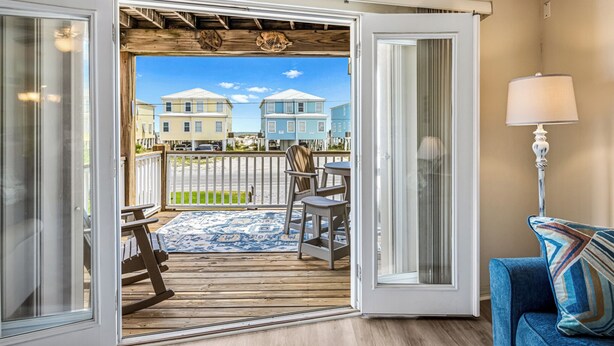 Puerta corredera abierta que da a una terraza de madera con vistas a casas, una mecedora, una mesa y una lámpara.