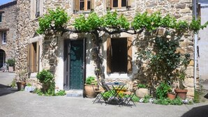 Un edificio de piedra con una puerta verde, dos ventanas con contraventanas de madera y macetas con plantas en la fachada.