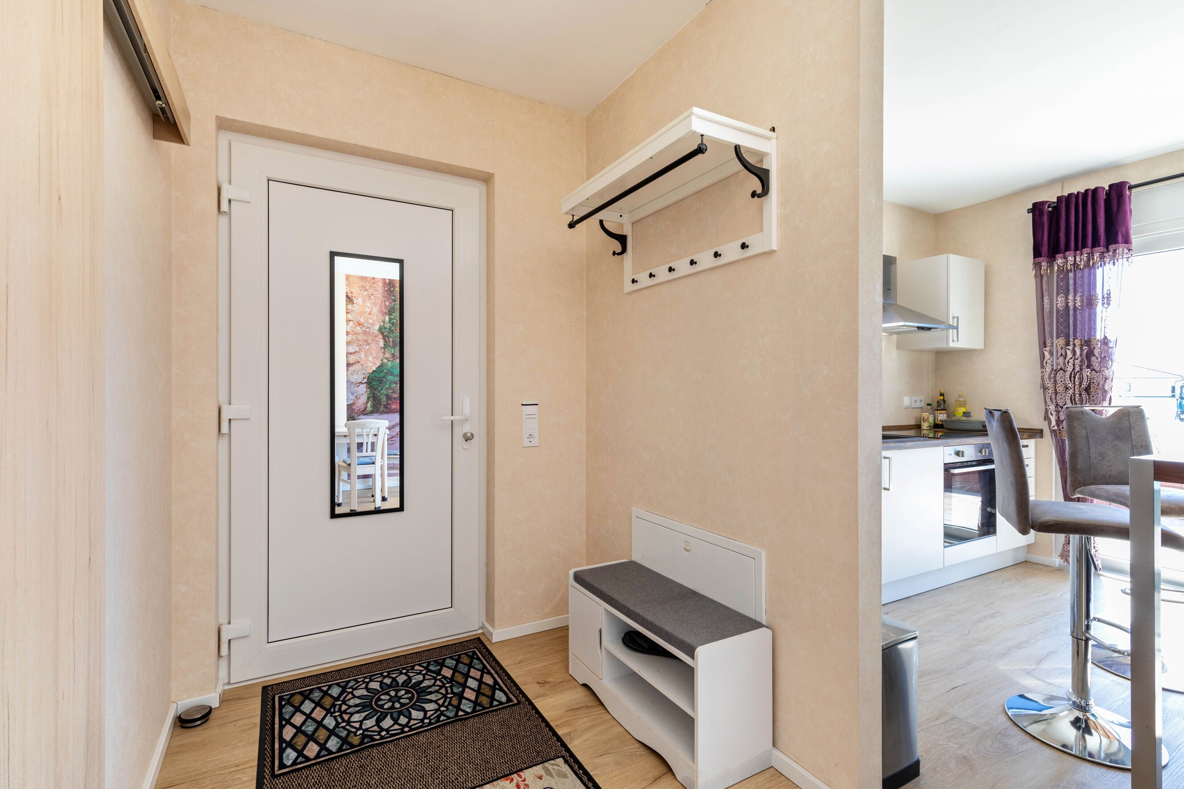 A hallway with a door, a small bench, a coat rack, and a kitchen area with a chair and a window.