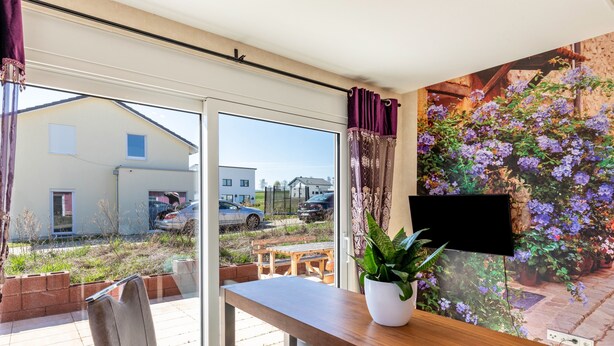 A room with a large window, a wooden table, a chair, a potted plant, and a wall-mounted TV. - 'Feel Free Apartment' holiday flat with private terrace and Wi-Fi (Rheinböllen)