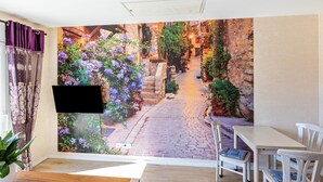 A room with a mural of a narrow street, a flat-screen TV, a dining table with chairs, and a potted plant. - 'Feel Free Apartment' holiday flat with private terrace and Wi-Fi (Rheinböllen)