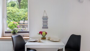 A dining area with a white table, two black chairs, a vase with flowers, and a window with a view of greenery.