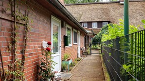 A narrow brick pathway leading to a house with a thatched roof, a metal fence, and potted plants.