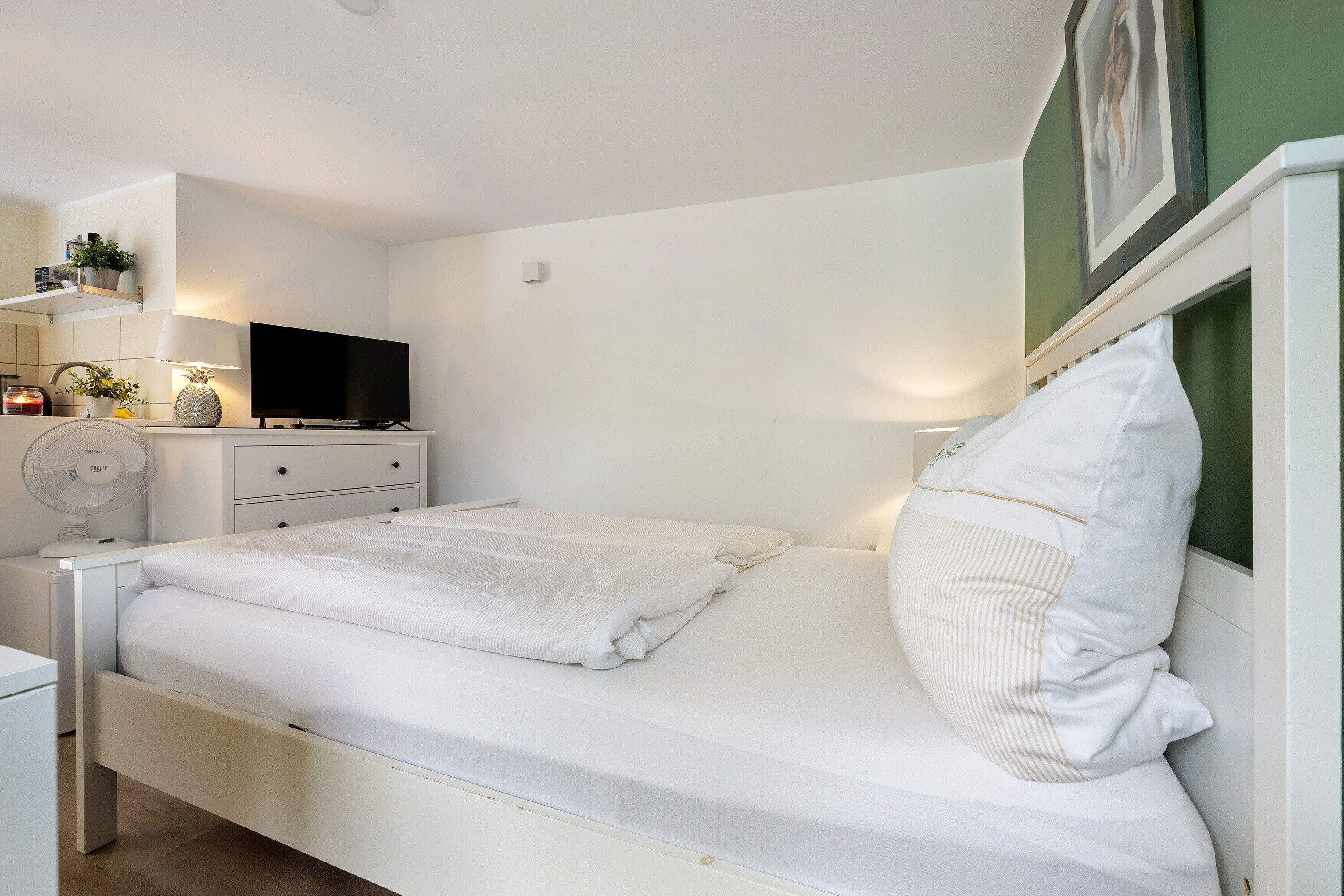 A neatly made bed with white linens, a bedside table with a lamp, a television on a stand, and a wall-mounted shelf with decorative items.