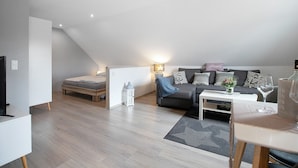 Una habitación moderna estilo loft con techo inclinado, un conjunto de sofás, una pequeña mesa de comedor y un televisor.