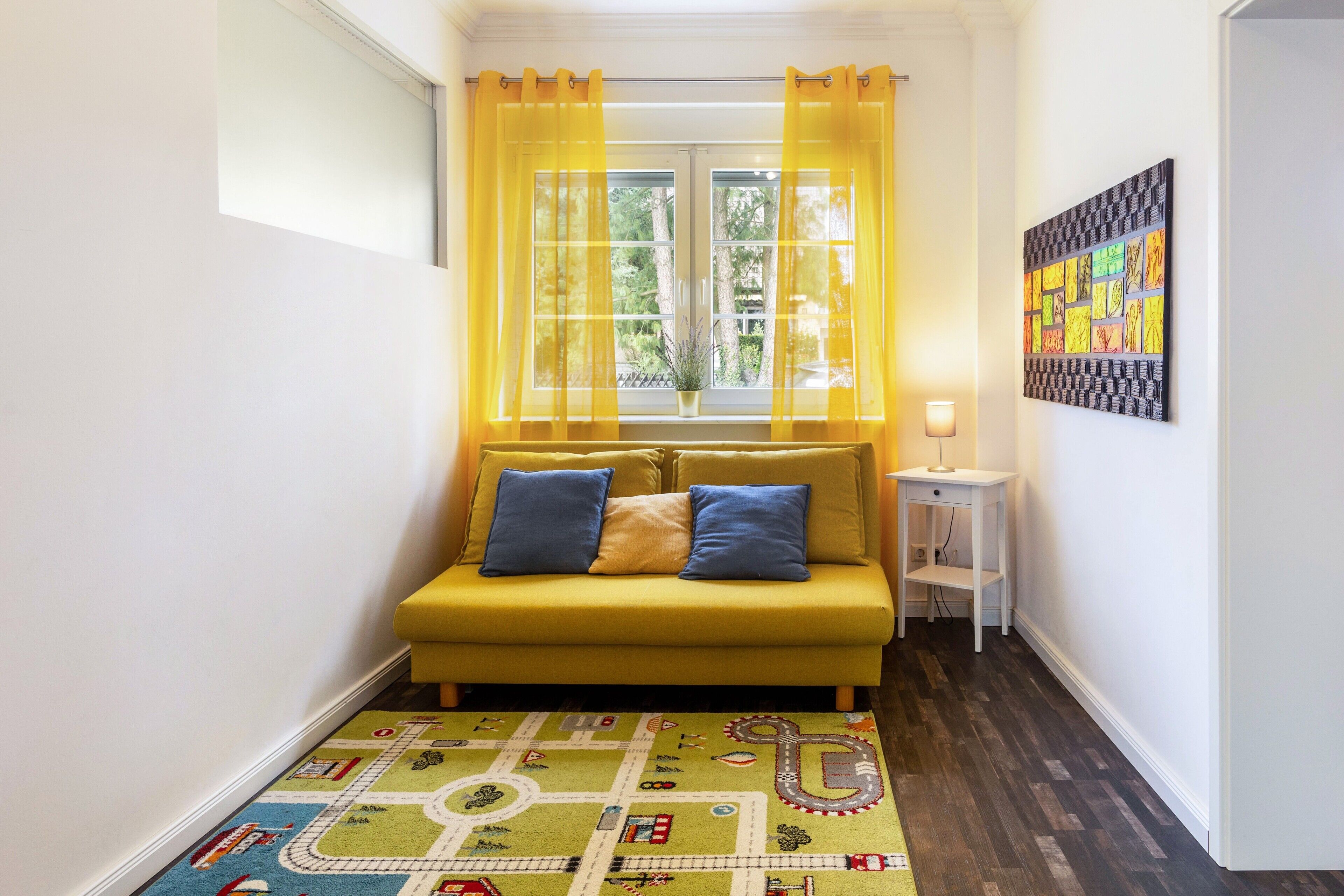 A room with a yellow sofa, a patterned rug, a window with yellow curtains, and a wall-mounted artwork.