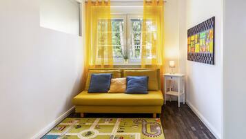A room with a yellow sofa, a patterned rug, a window with yellow curtains, and a wall-mounted artwork.
