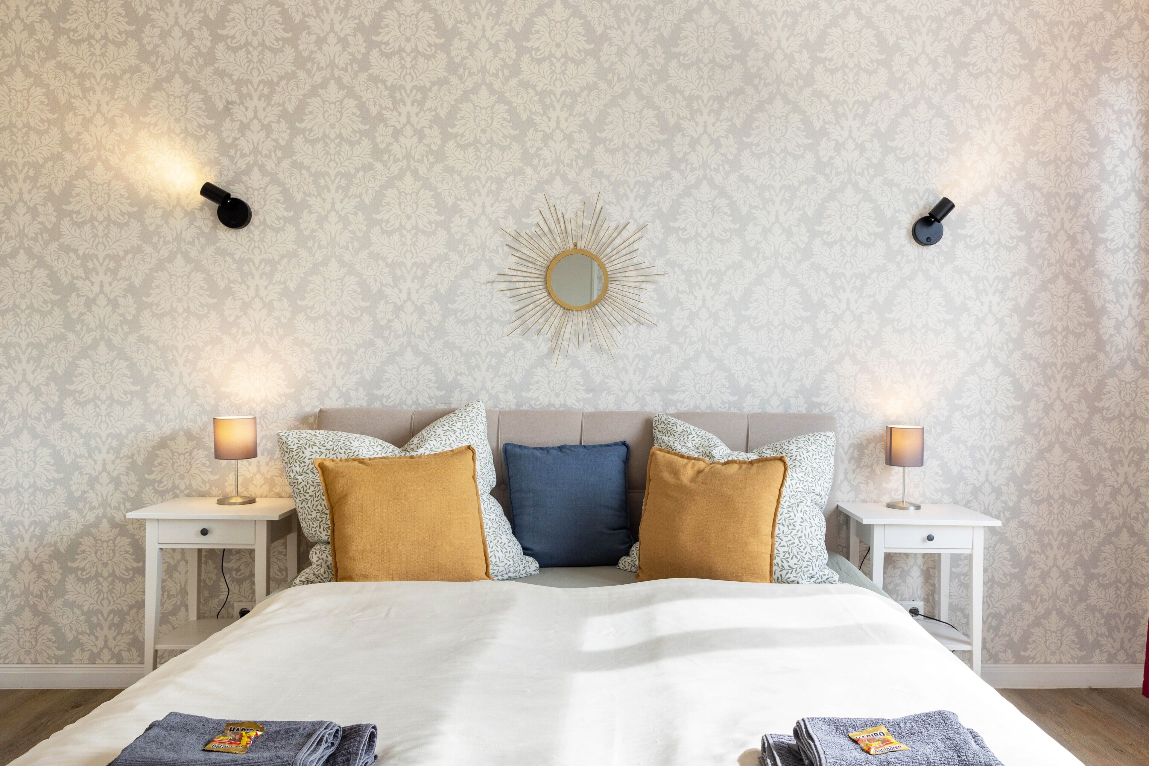 A neatly made bed with a white duvet, two bedside tables with lamps, patterned wallpaper, and a sunburst mirror on the wall.
