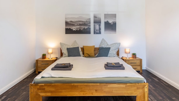 Un lit bien fait, avec une tête de lit en bois, deux tables de chevet avec des lampes et trois tableaux accrochés au mur.