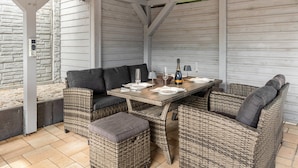 Un patio couvert aménagé avec des meubles en osier, une table dressée pour le repas et un mur de pierre en arrière-plan.