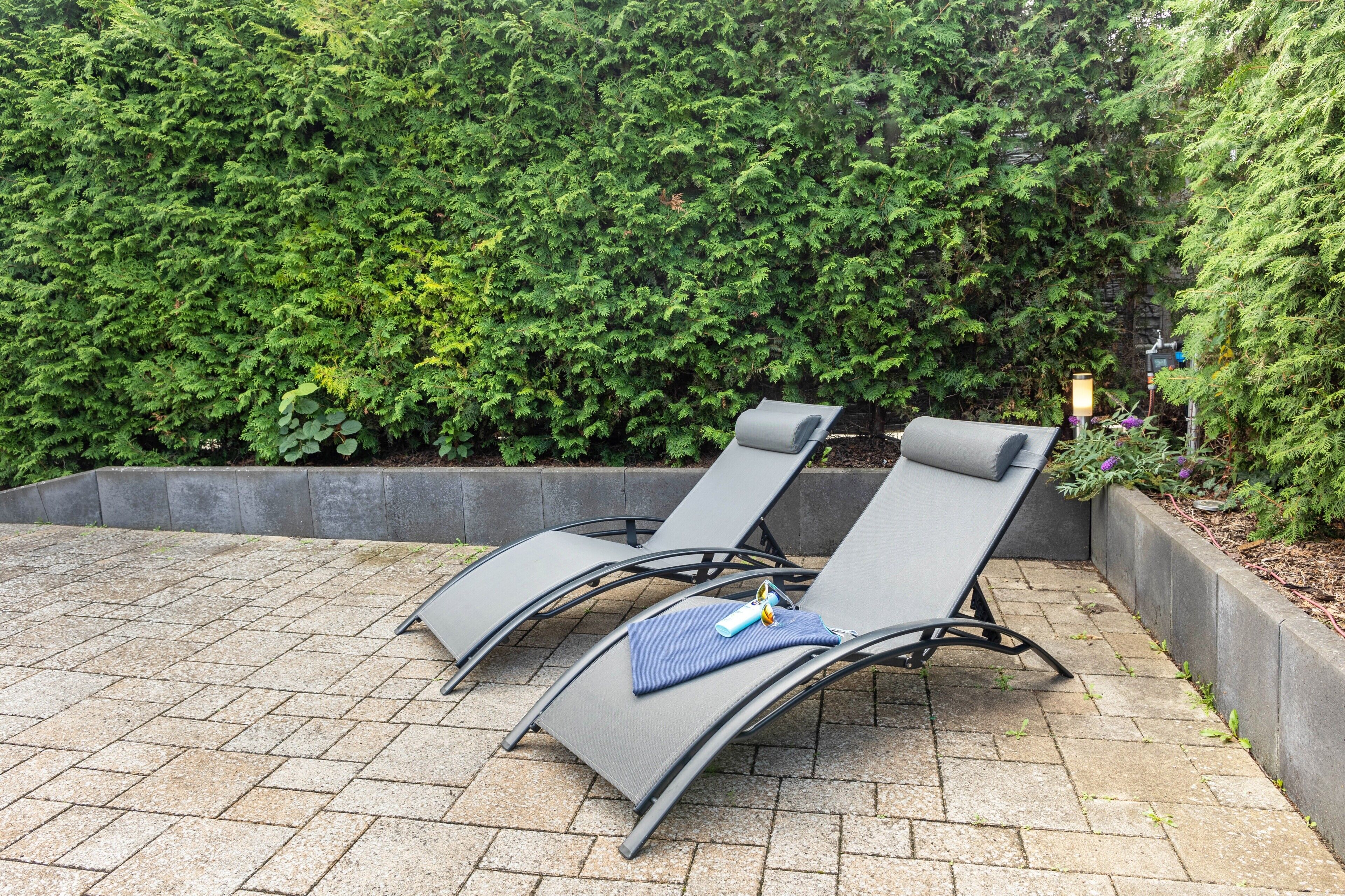 Two lounge chairs with a cushion and a bottle, set on a paved patio with a hedge in the background.