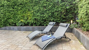 Two lounge chairs with a cushion and a bottle, set on a paved patio with a hedge in the background.