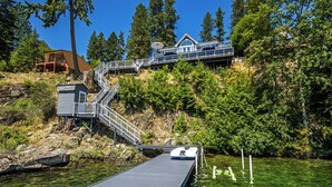 A wooden dock extending into a clear lake, surrounded by lush greenery and a hillside with a modern house.