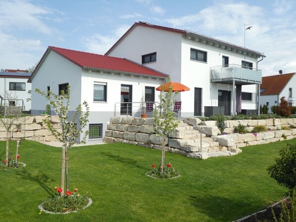 Une maison moderne à deux étages, avec un toit rouge, des marches en pierre et une pelouse bien entretenue agrémentée de fleurs.