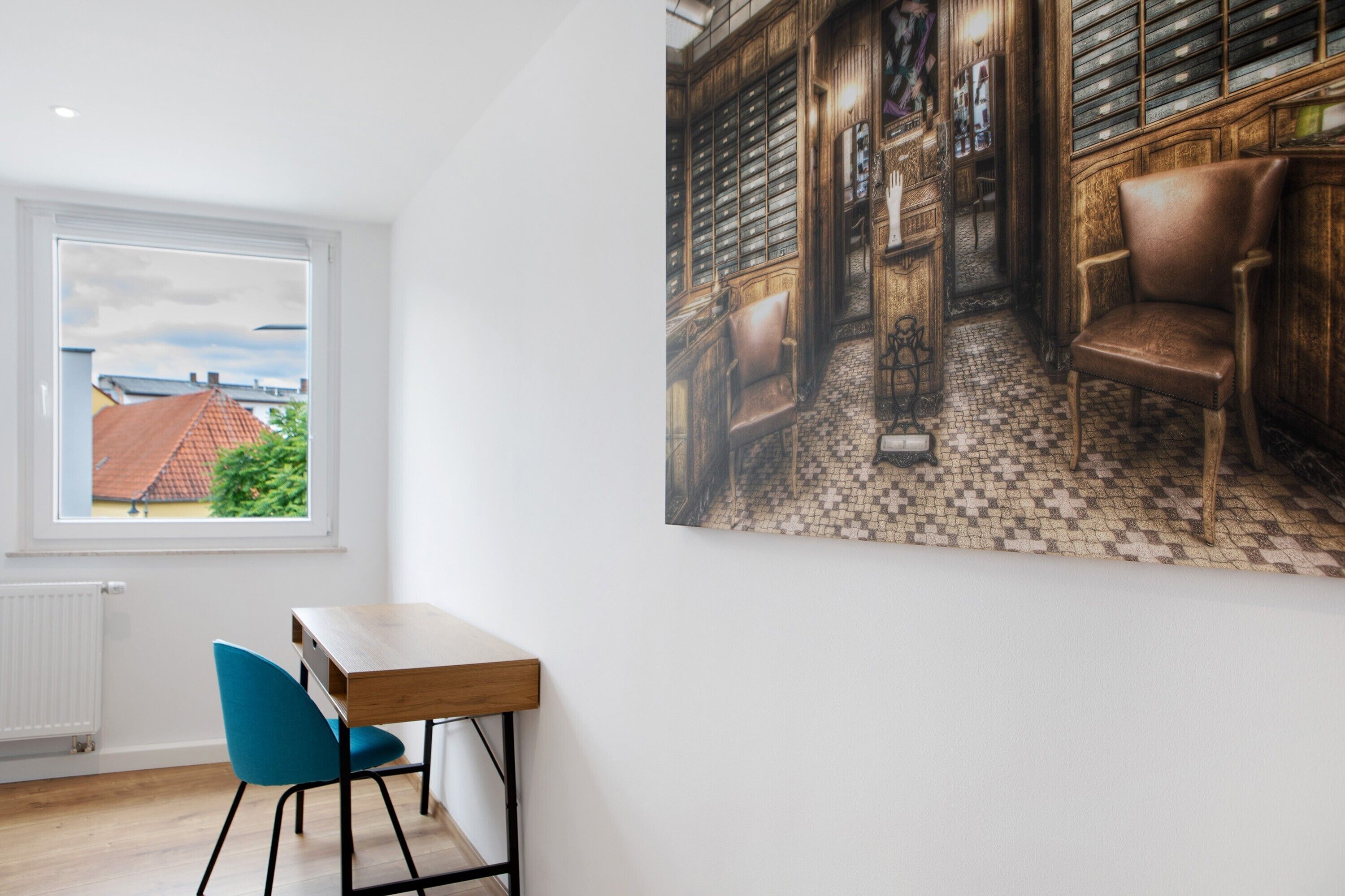 A room with a window, a desk, a chair, and a large framed picture on the wall.
