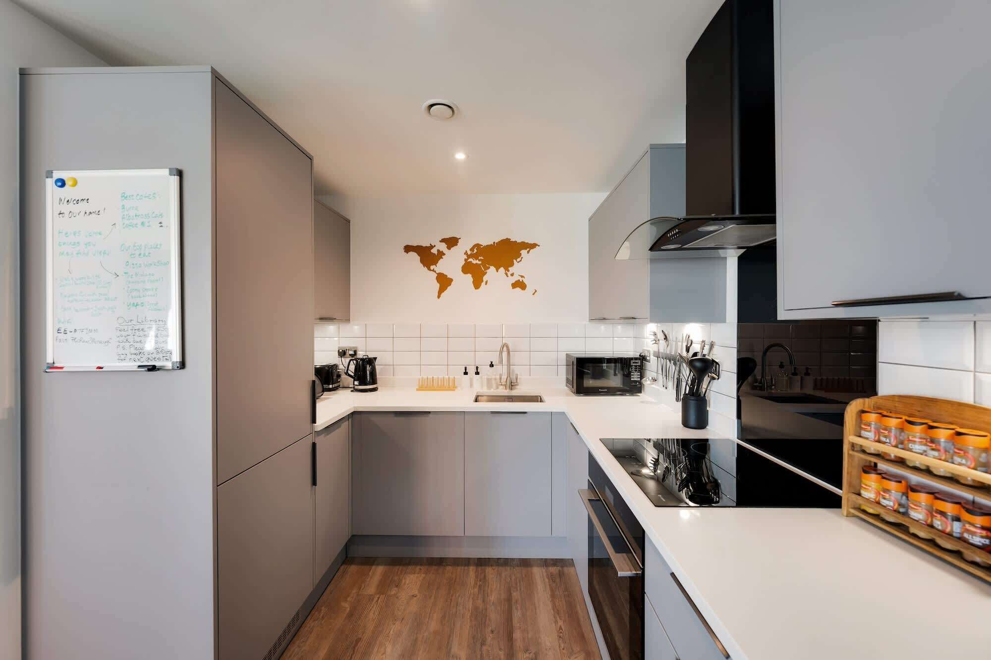 Une cuisine moderne avec un réfrigérateur blanc, un four à micro-ondes, une machine à café et un évier. Un grand planisphère est accroché au mur, et on y trouve également un petit îlot central doté d’un évier et d’un plan de travail.