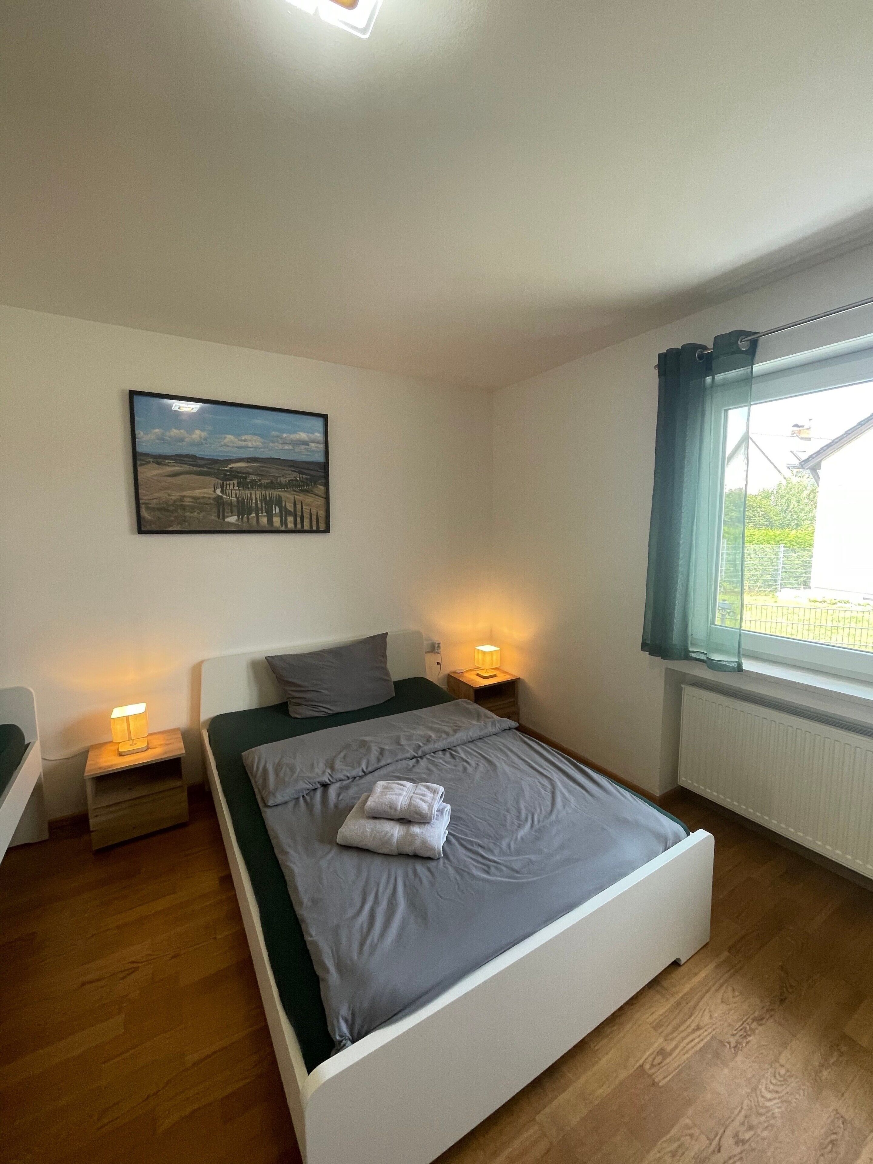 Une chambre à coucher avec un lit, deux tables de chevet avec des lampes, un tableau accroché au mur et une fenêtre avec des rideaux.