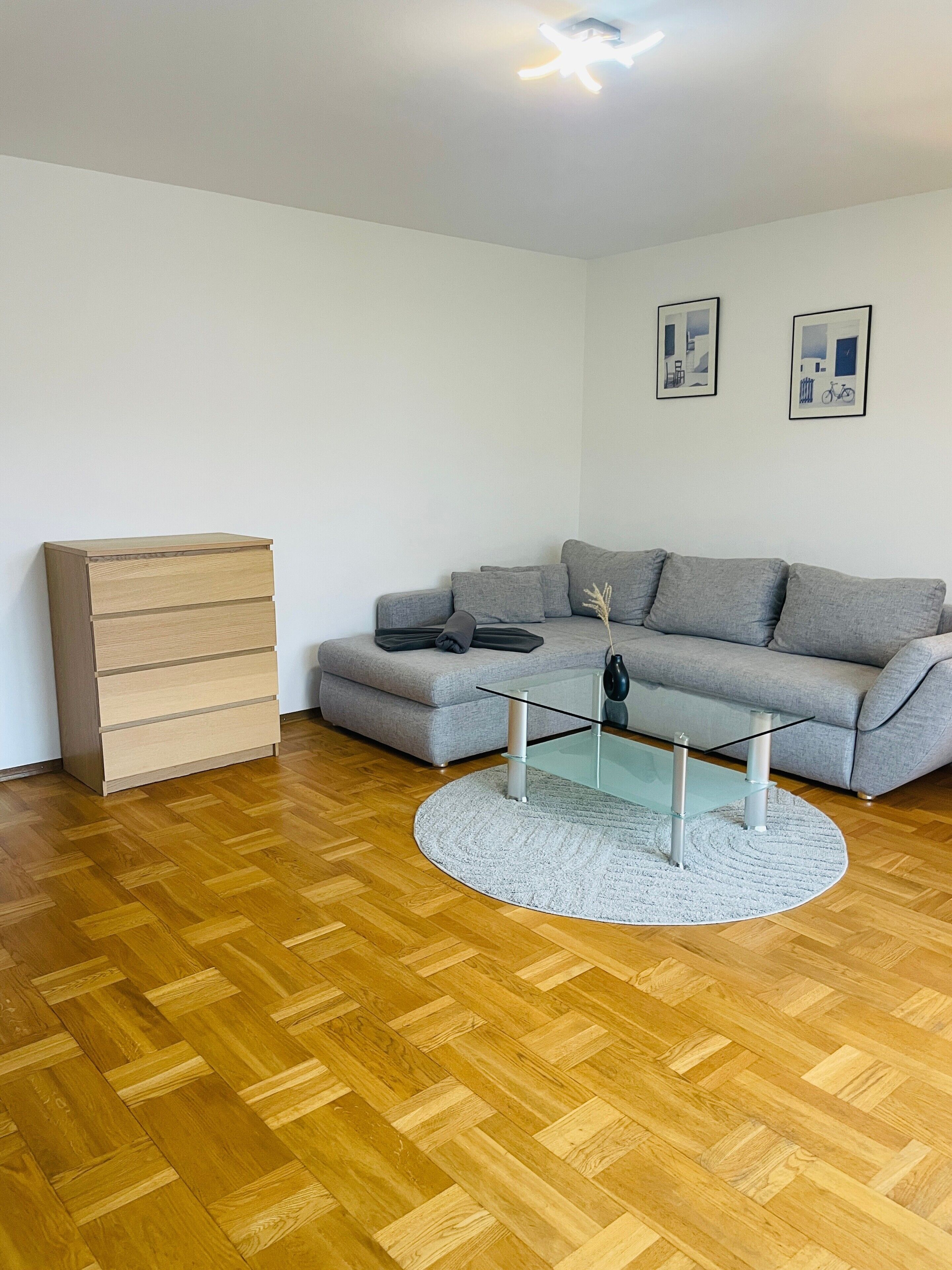 A living room with a grey sofa, a glass coffee table, a wooden chest of drawers, and a herringbone wooden floor.