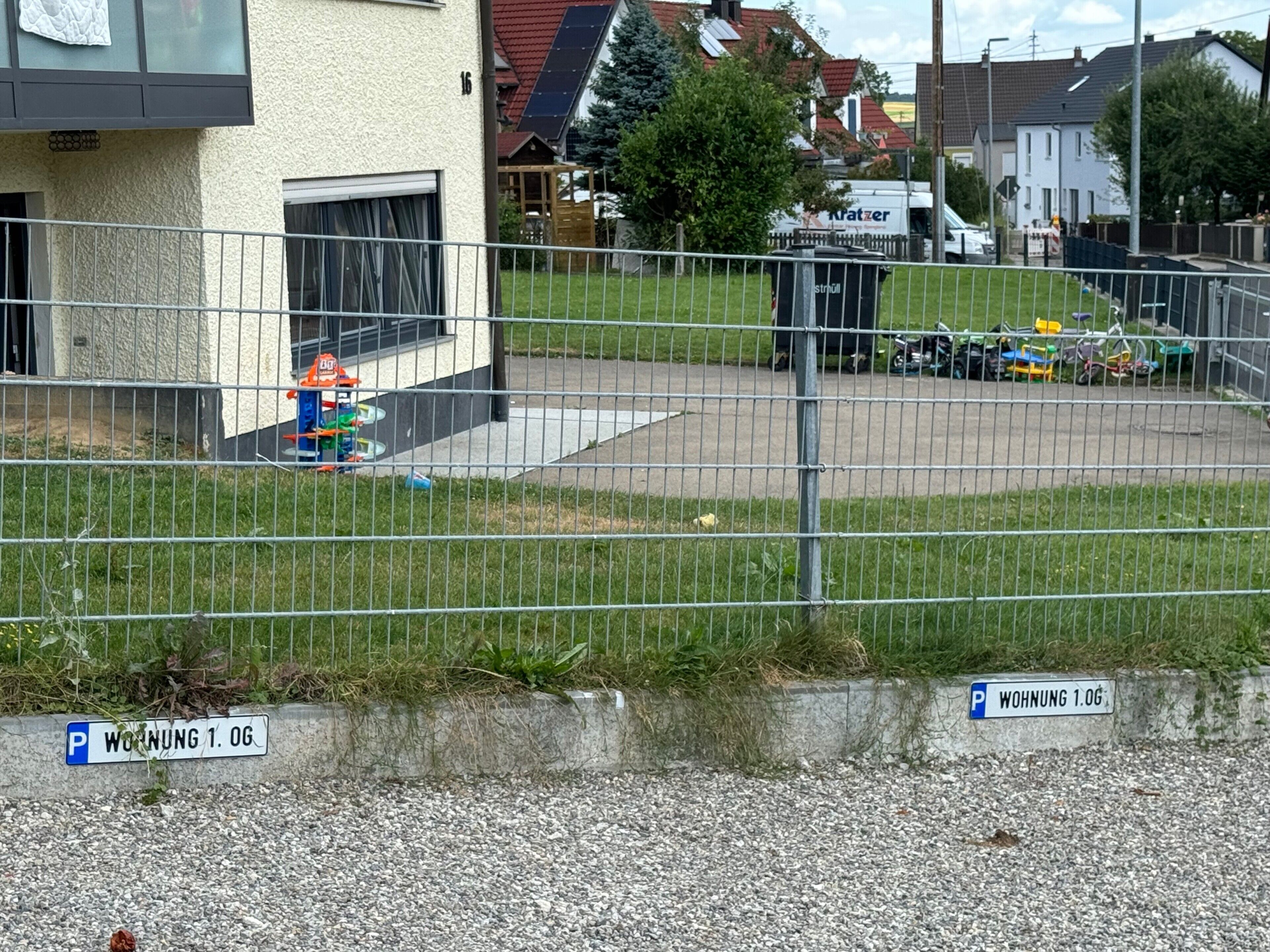 Ein eingezäunter Parkbereich mit Kiesbefestigung, ein Gebäude mit weißer Fassade und ein sichtbarer Hydrant.