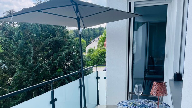 Un balcón con mesa y sillas, barandilla de vidrio y vistas a árboles y edificios.