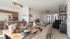 Une cuisine moderne avec un îlot central en bois, un coin repas avec une longue table dressée pour six personnes, et un canapé en cuir noir dans l’espace salon.