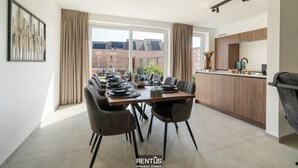 Un espace de repas moderne avec une longue table dressée pour six personnes, une cuisine dotée de meubles en bois et de grandes fenêtres offrant une vue sur le paysage urbain.