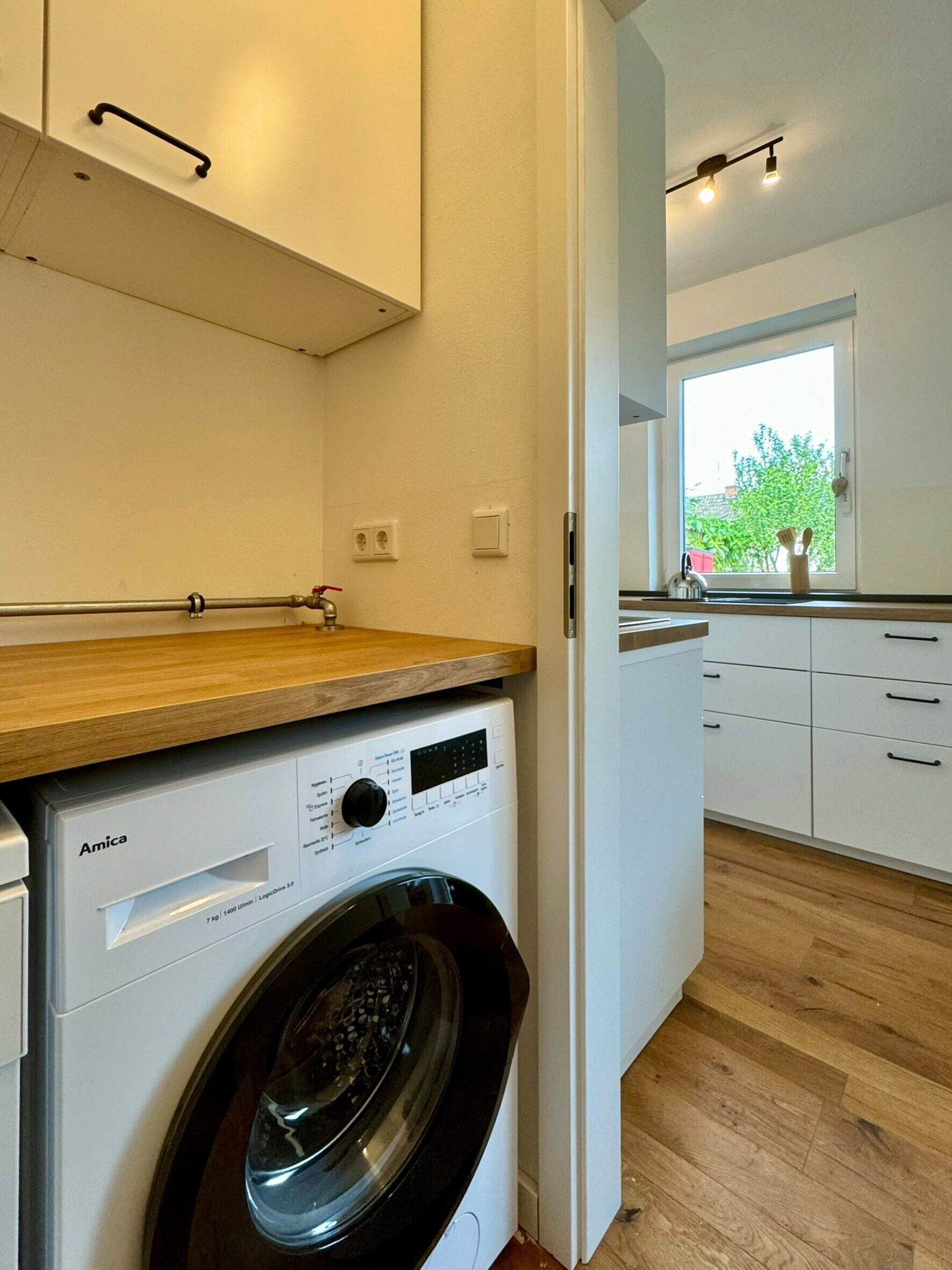 A compact laundry area with a front-loading Amica washing machine, wooden countertop, white cabinets, and a window with a view of greenery.
