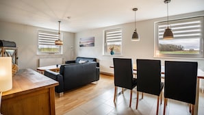 A modern living room with a sofa, dining table, chairs, pendant lights, and a view through large windows. - Apartment 'Strandläufer' with Shared Garden and Wi-Fi (Westerholz)