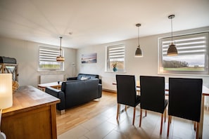 Una sala de estar moderna con sofá, mesa de comedor, sillas, lámparas colgantes y vistas amplias a través de grandes ventanales.