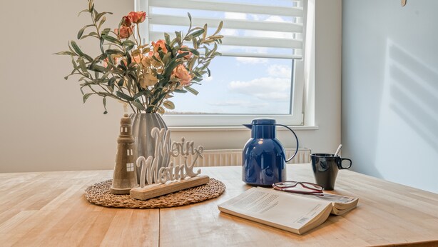 Une table en bois sur laquelle se trouvent un vase de fleurs, une bouilloire bleue, une tasse et une paire de lunettes.
