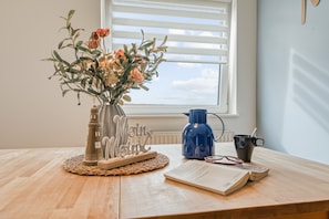 Une table en bois sur laquelle se trouvent un vase de fleurs, une bouilloire bleue, une tasse et une paire de lunettes.