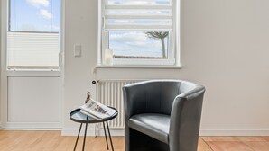 Une pièce comprenant un fauteuil en cuir gris, une petite table avec un magazine et deux fenêtres équipées de stores.
