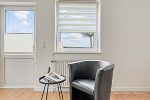 A room with a grey leather chair, a small table with a magazine, and two windows with blinds. - Holiday Apartment 'Ankerplatz' with Private Terrace, Shared Garden & Wi-Fi (Westerholz)