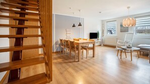 Un intérieur moderne avec un escalier en bois, un coin repas comprenant une table et des chaises, des luminaires suspendus et une vue dégagée grâce à de grandes fenêtres.