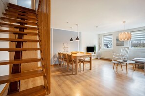 A modern interior with a wooden staircase, a dining area with a table and chairs, pendant lights, and a view through large windows.