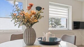 A dining table with a vase of flowers, a bowl with teapots, a magazine, and a window with blinds.
