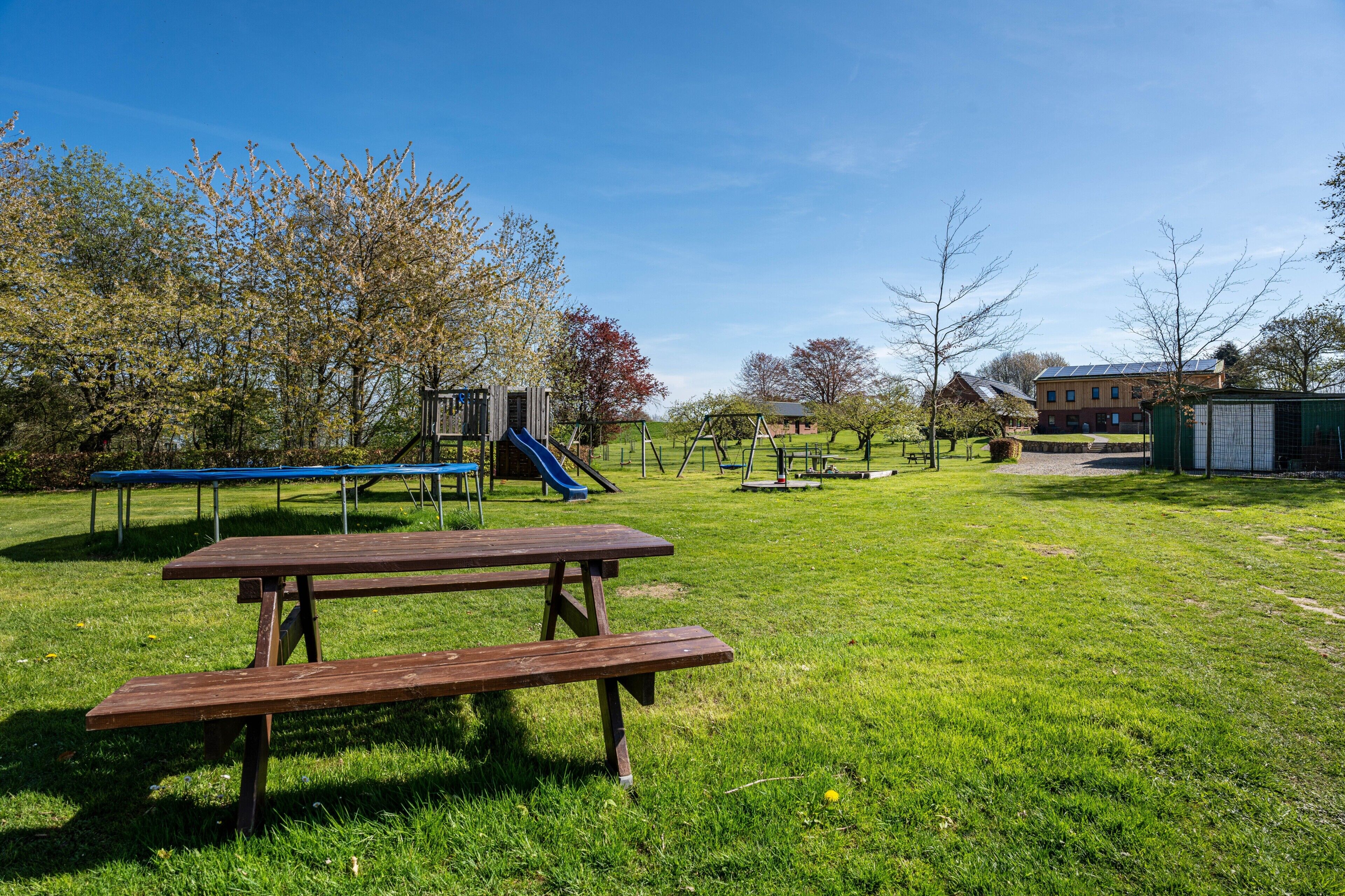 A picnic table and playground equipment in a grassy park area with trees and a building in the background.