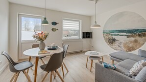 A modern living room with a dining area, a grey sofa, and a large wall mural of a beach scene. - Apartment 'Küstenkind' with Private Terrace, Private Garden and Wi-Fi (Westerholz)