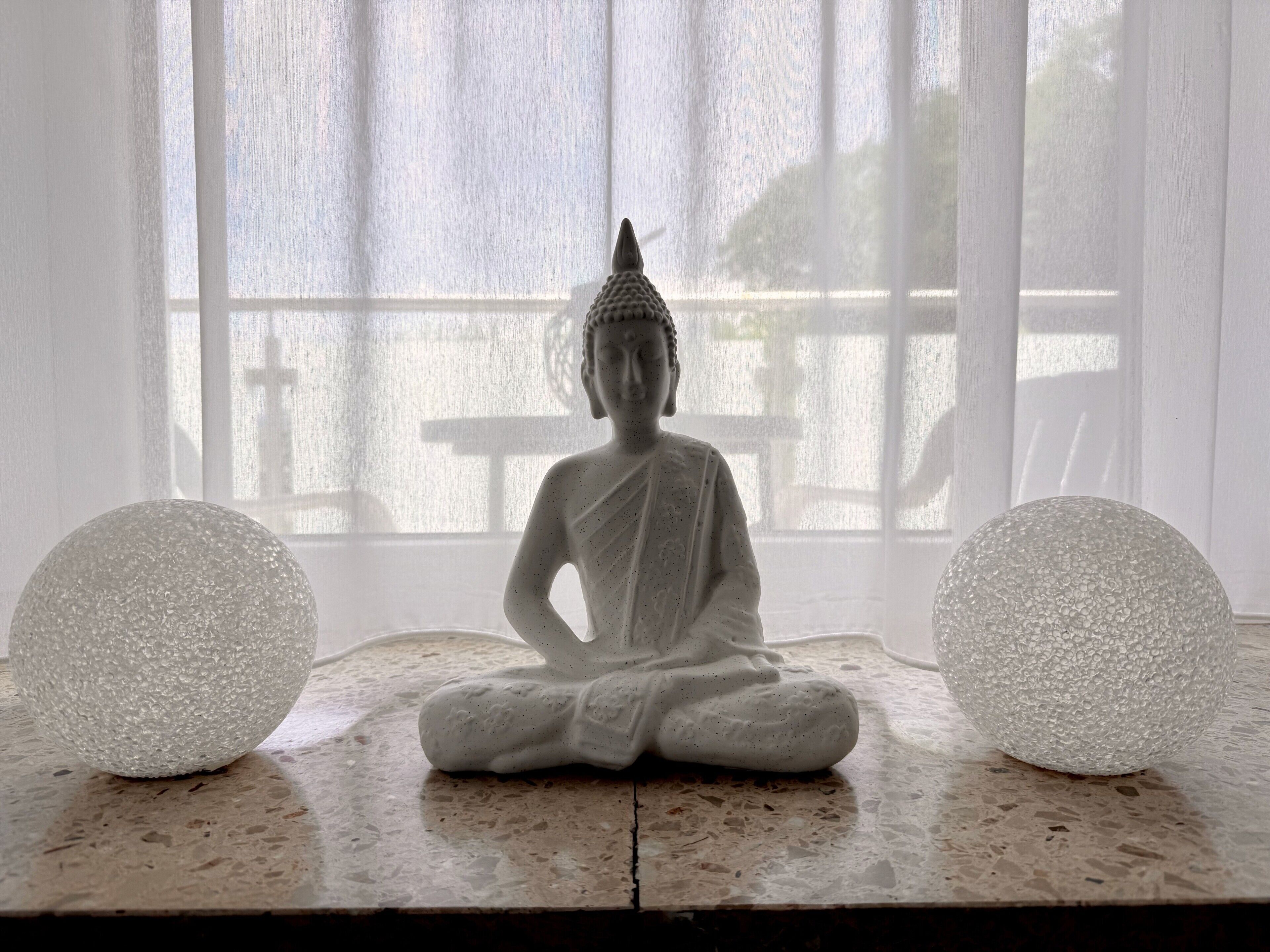 Una estatua sentada en posición meditativa, ubicada entre dos objetos esféricos con textura, con una ventana al fondo.