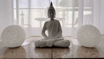 Una estatua sentada en posición meditativa, ubicada entre dos objetos esféricos con textura, con una ventana al fondo.