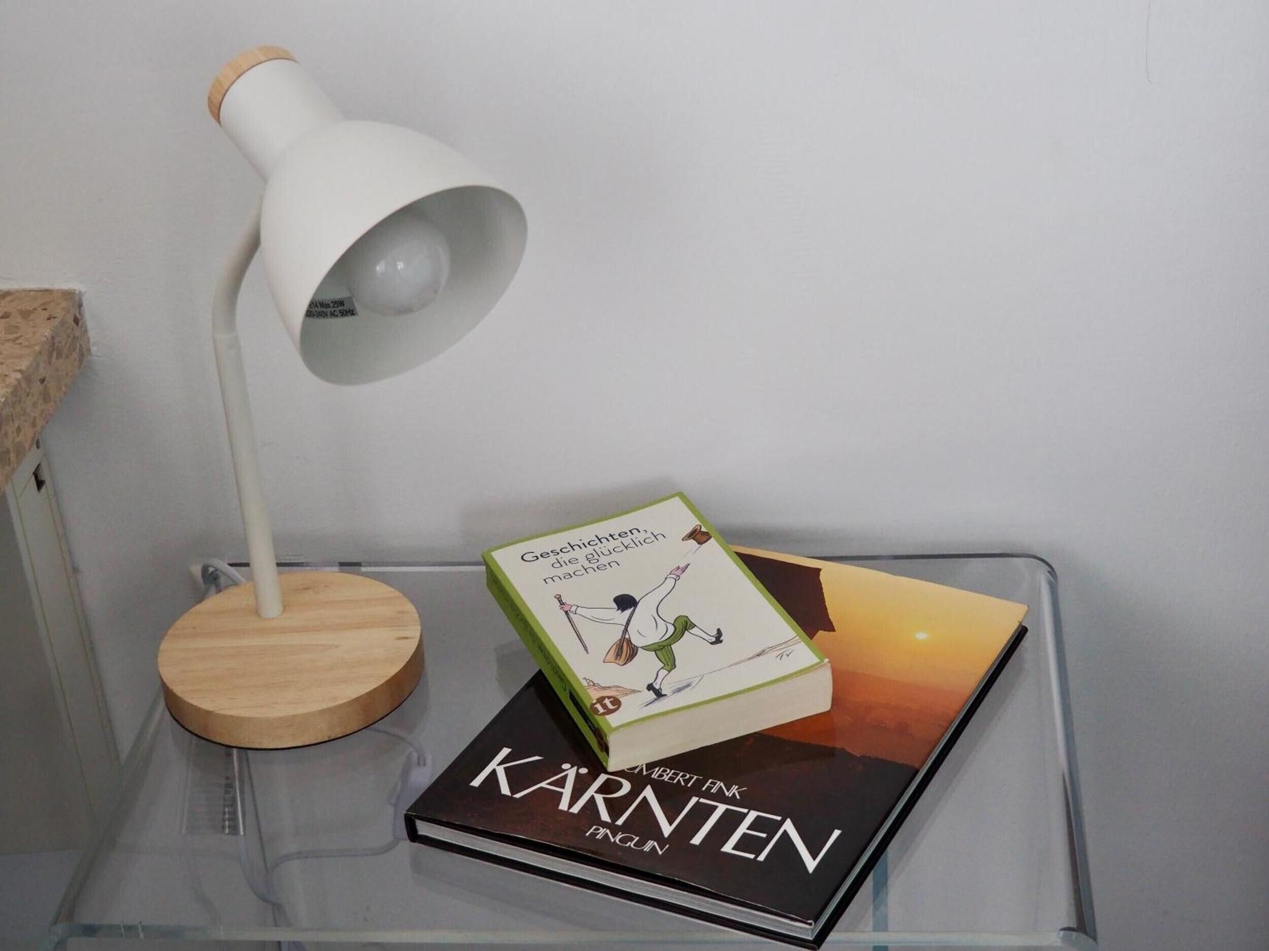 A book titled 'Kärnten' by Albert Fink on a clear glass table, a white lamp with a wooden base, and a partial view of a textured wall.