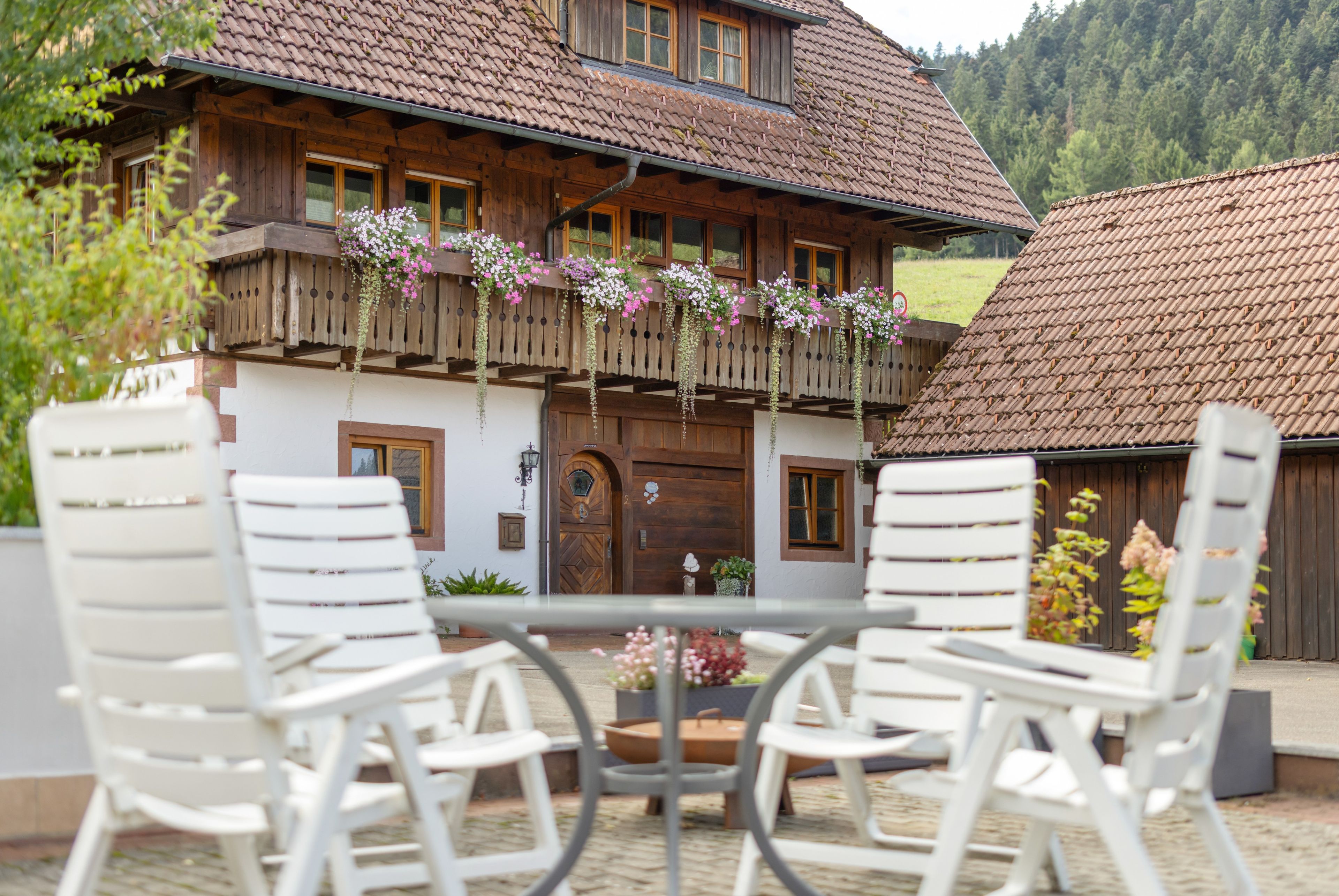 A wooden chalet with a balcony adorned with flowers, a wooden door, and a view of a forested hillside.