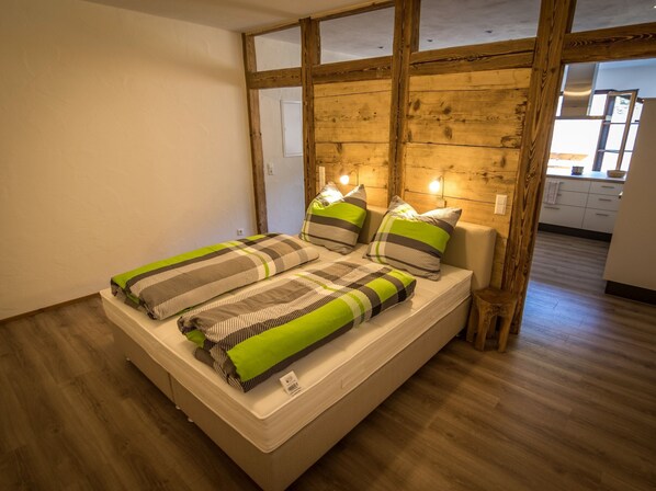 A single bed with striped bedding and pillows, wooden beams on the ceiling, and a visible kitchen area in the background.