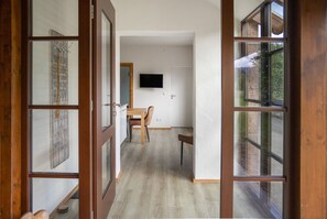 Un pasillo con piso de madera, un comedor con mesa y sillas, un televisor montado en la pared y un ventanal con vista a follaje.