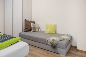 Una habitación pequeña con cama individual, un sofá cama gris, una mesita de noche blanca y una almohada verde.